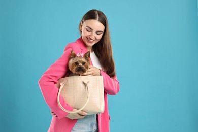 Beautiful young woman carrying cute Yorkshire Terrier dog in bag on light blue background, space for text