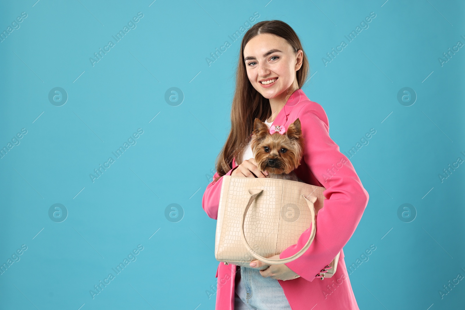 Photo of Beautiful young woman carrying cute Yorkshire Terrier dog in bag on light blue background, space for text