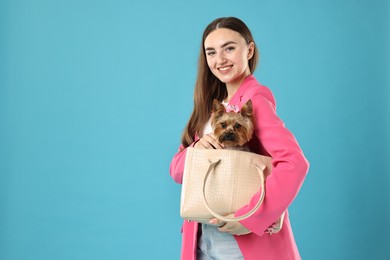 Beautiful young woman carrying cute Yorkshire Terrier dog in bag on light blue background, space for text