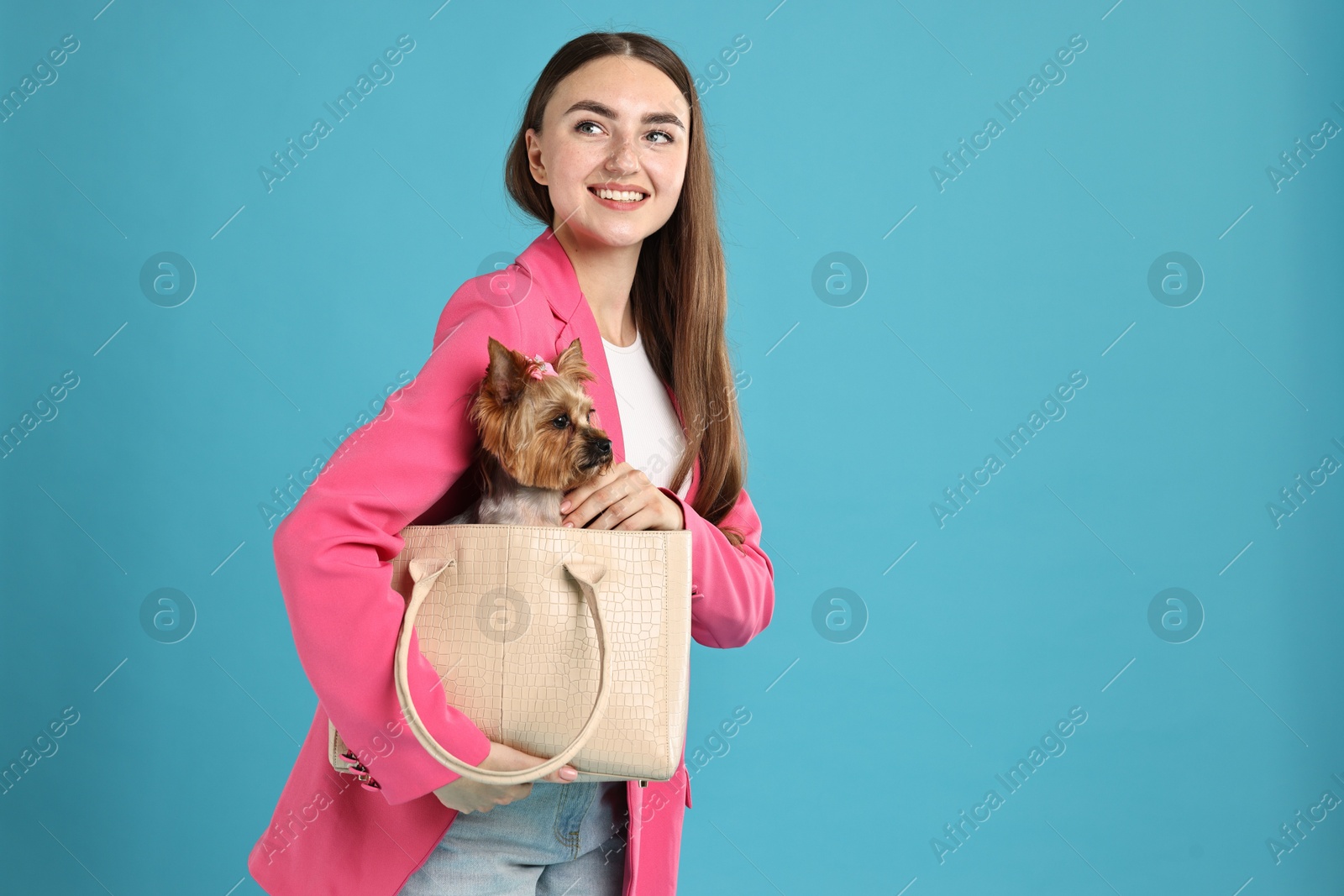 Photo of Beautiful young woman carrying cute Yorkshire Terrier dog in bag on light blue background, space for text