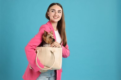 Photo of Beautiful young woman carrying cute Yorkshire Terrier dog in bag on light blue background, space for text