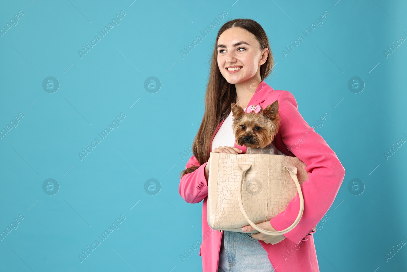 Photo of Beautiful young woman carrying cute Yorkshire Terrier dog in bag on light blue background, space for text