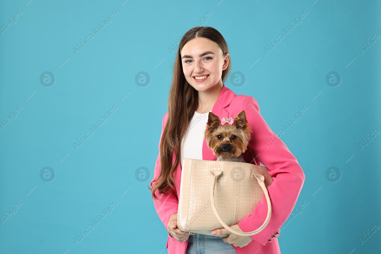 Photo of Beautiful young woman carrying cute Yorkshire Terrier dog in bag on light blue background