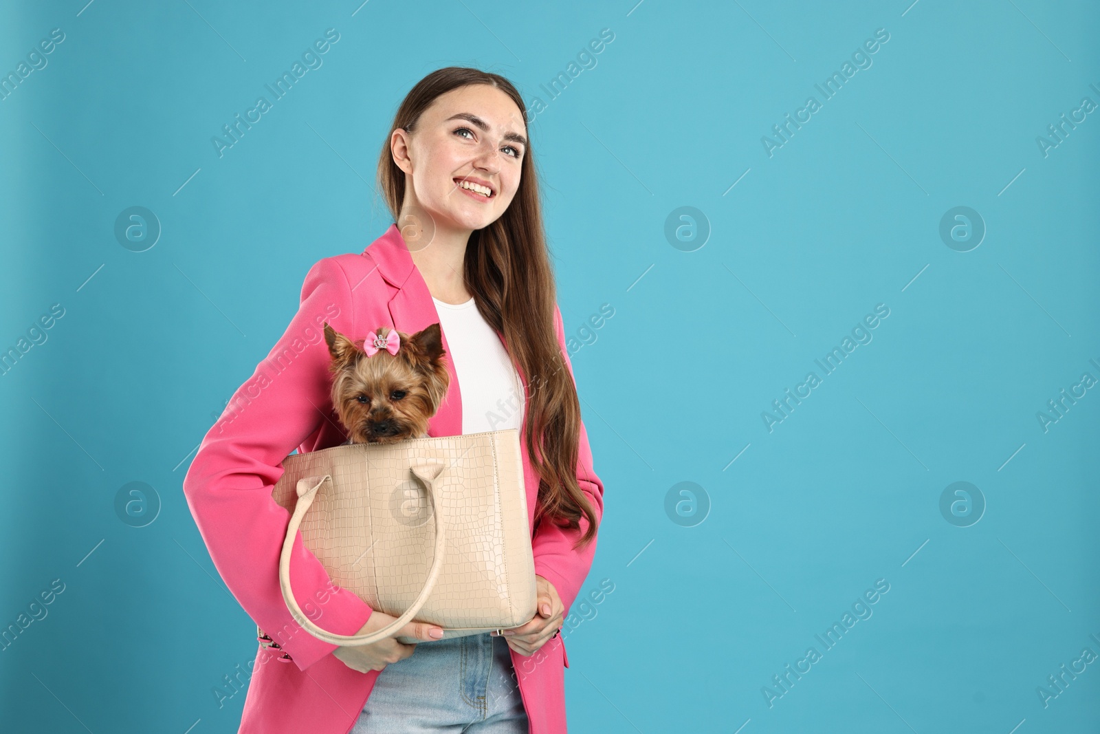 Photo of Beautiful young woman carrying cute Yorkshire Terrier dog in bag on light blue background, space for text