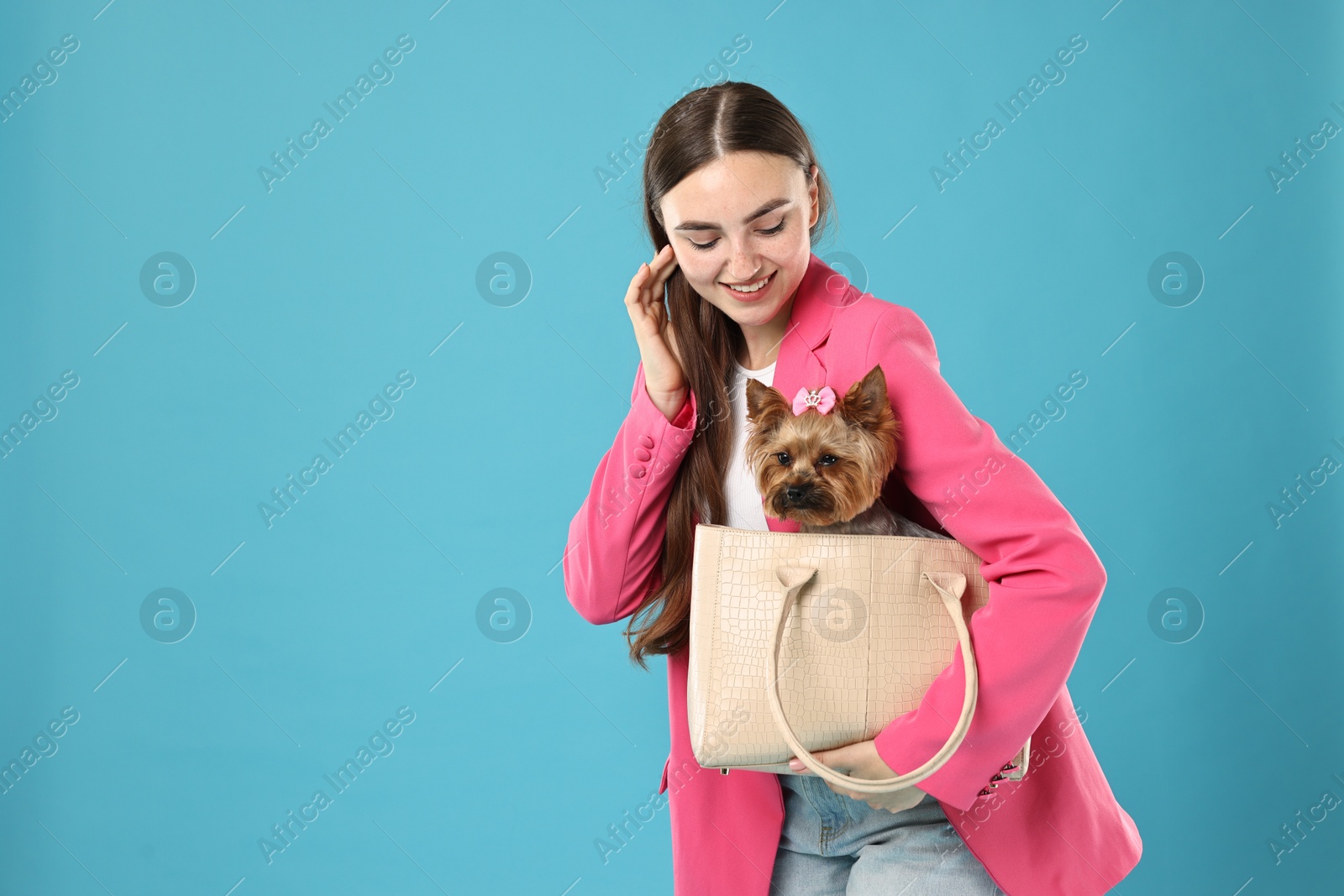 Photo of Beautiful young woman carrying cute Yorkshire Terrier dog in bag on light blue background, space for text