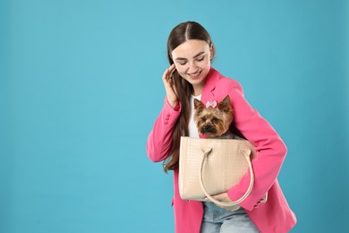 Photo of Beautiful young woman carrying cute Yorkshire Terrier dog in bag on light blue background, space for text