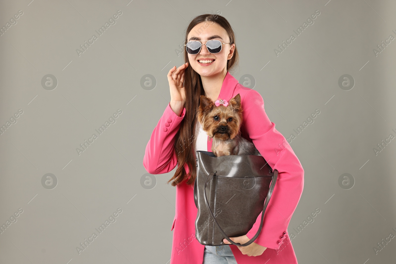 Photo of Woman in sunglasses carrying cute Yorkshire Terrier dog in bag on grey background