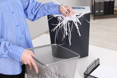 Photo of Woman putting shredded paper strips into trash bin at white table in office, closeup