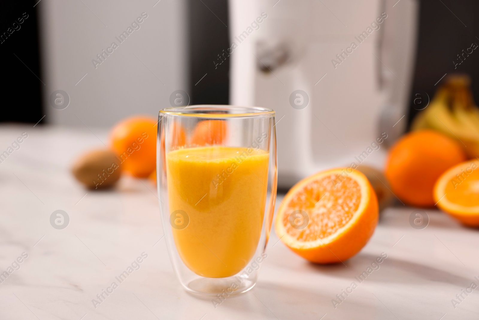 Photo of Glass of fresh juice, modern juicer and fruits on white marble table in kitchen, selective focus