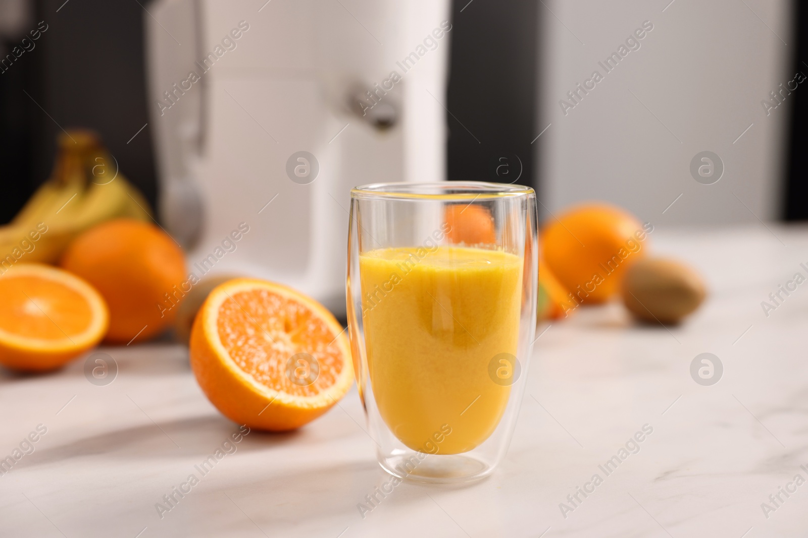 Photo of Glass of fresh juice, modern juicer and fruits on white marble table in kitchen, selective focus