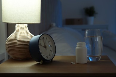 Photo of Insomnia treatment. Glass of water, pills and alarm clock on bedside table in bedroom at night