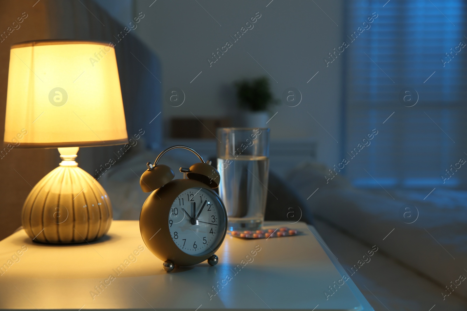 Photo of Insomnia treatment. Alarm clock, glass of water and pills on bedside table in bedroom at night