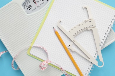 Photo of Body fat caliper, measuring tape, scale and notebook on light blue background, flat lay