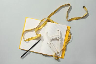 Photo of Body fat caliper, measuring tape and notebook on grey background, flat lay