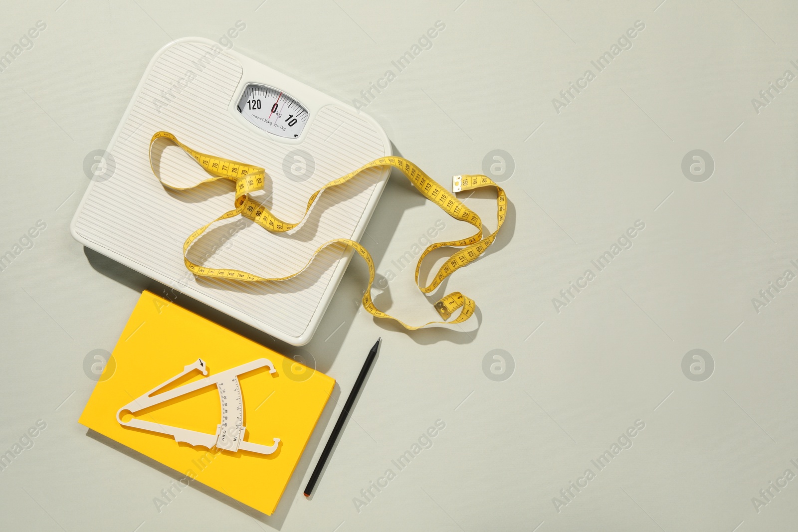 Photo of Body fat caliper, measuring tape, scale and notebook on light grey background, flat lay. Space for text