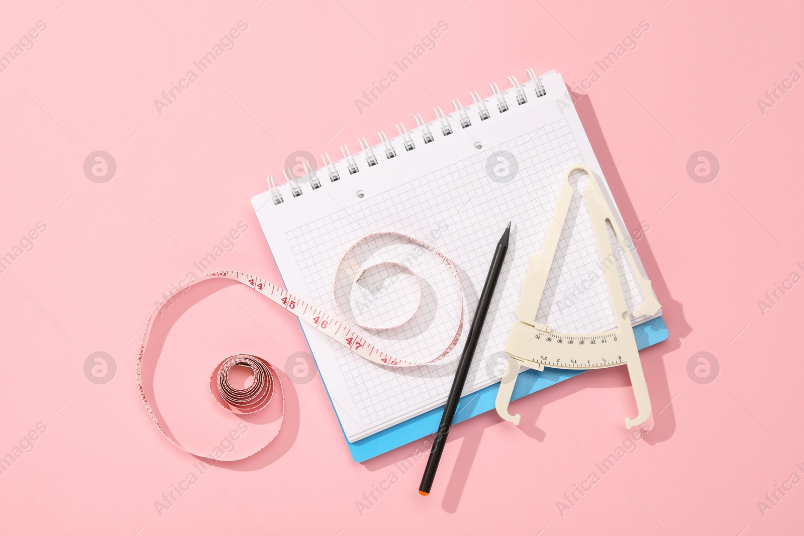 Photo of Body fat caliper, measuring tape, notebook and pencil on pink background, flat lay