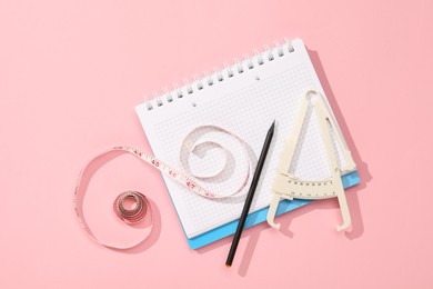 Photo of Body fat caliper, measuring tape, notebook and pencil on pink background, flat lay