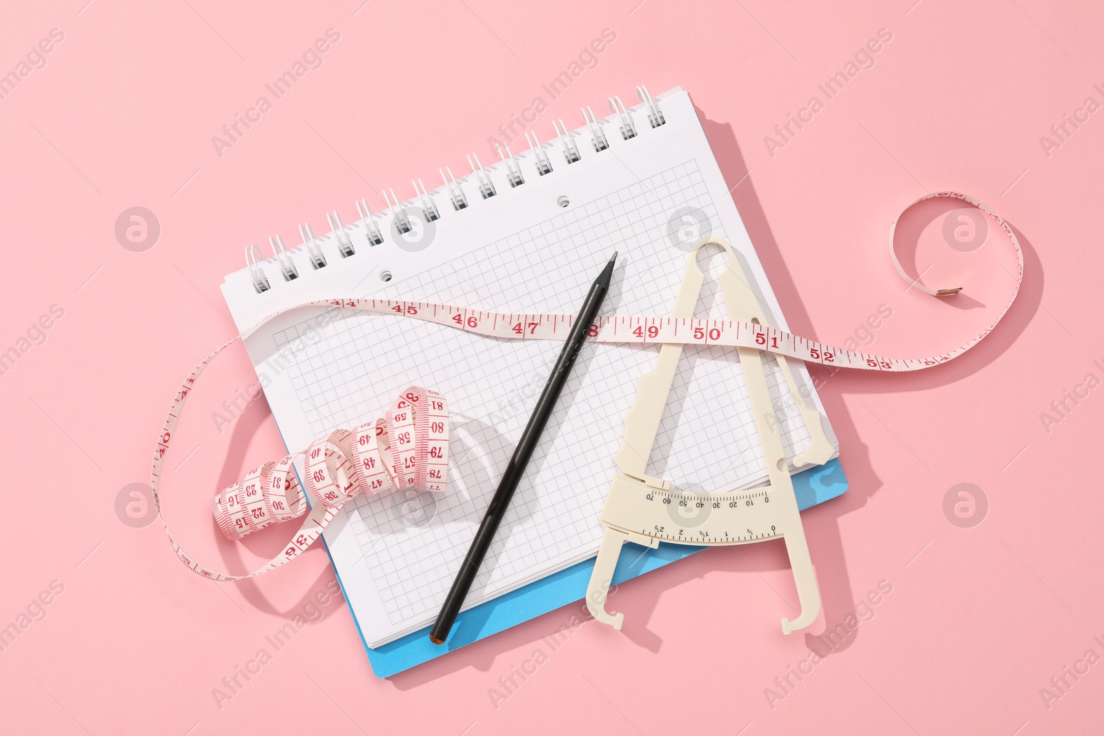 Photo of Body fat caliper, measuring tape, notebook and pencil on pink background, flat lay