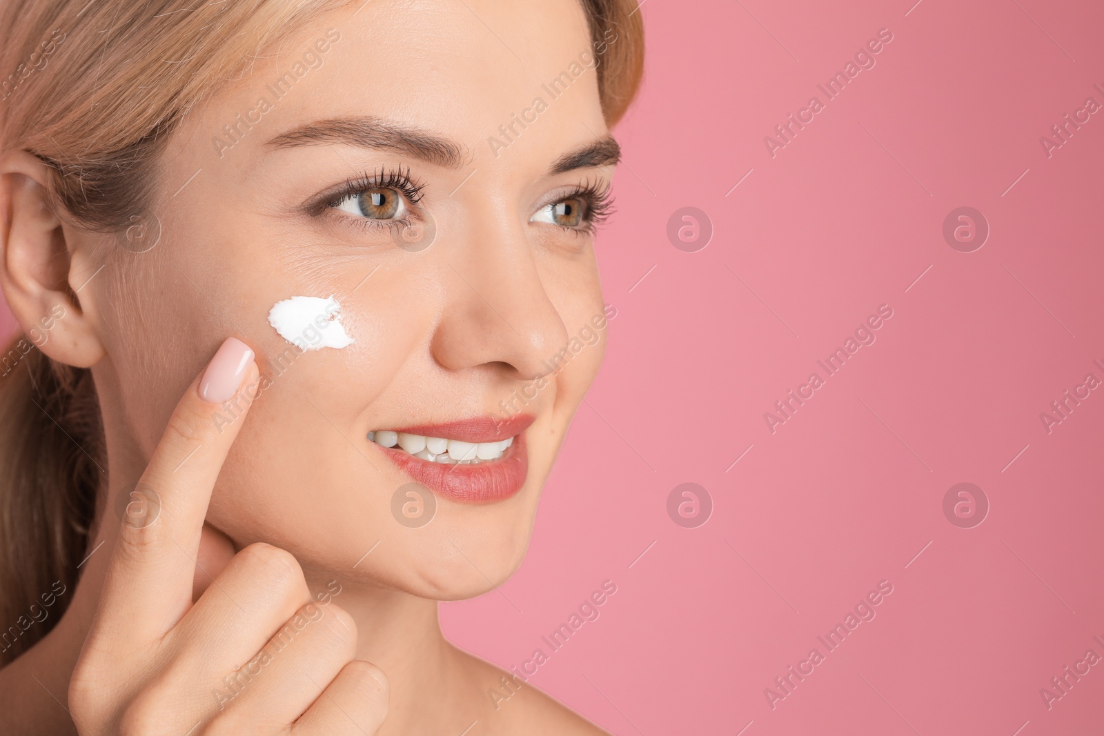 Photo of Beautiful woman applying cream on her face against pink background, closeup. Space for text