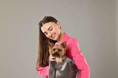 Beautiful young woman carrying cute Yorkshire Terrier dog in bag on grey background