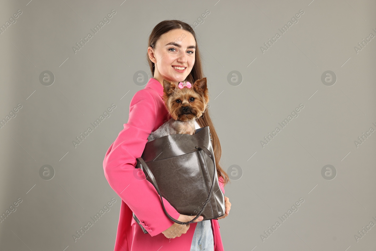 Photo of Beautiful young woman carrying cute Yorkshire Terrier dog in bag on grey background, space for text