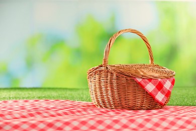 Photo of One picnic wicker basket with checkered napkin and blanket on grass against blurred green background. Space for text