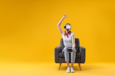 Photo of Happy woman with virtual reality headset and controller sitting in armchair on yellow background, space for text