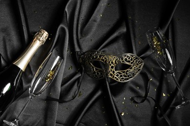 Beautiful carnival mask, bottle of champagne and glasses on black silk fabric, flat lay