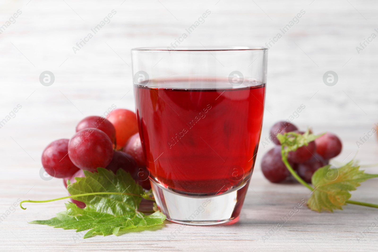 Photo of Tasty grape juice in glass, leaves and berries on light wooden table, closeup