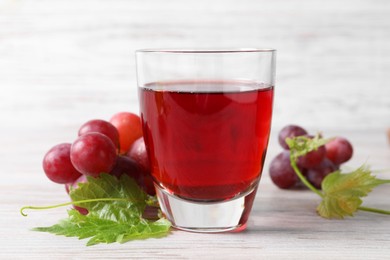 Photo of Tasty grape juice in glass, leaves and berries on light wooden table, closeup