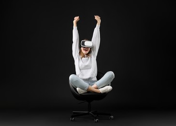 Happy woman with virtual reality headset sitting on chair against black background