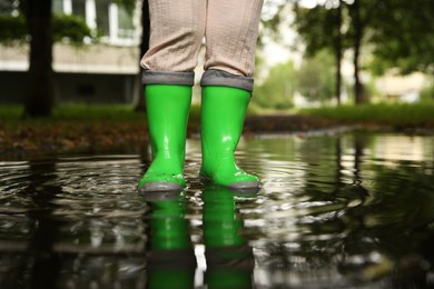 Photo of Little girl wearing green rubber boots standing in puddle outdoors, closeup