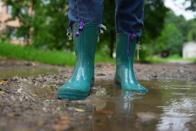 Photo of Woman wearing turquoise rubber boots walking in puddle outdoors, closeup