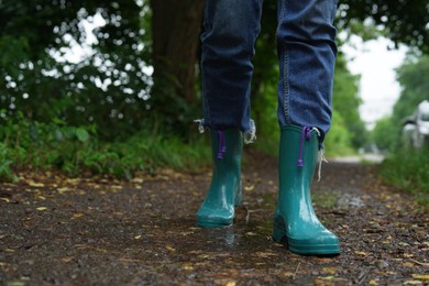 Woman in turquoise rubber boots walking on wet road, closeup. Space for text