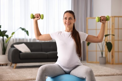 Beautiful pregnant woman with dumbbells doing exercises on fitball at home