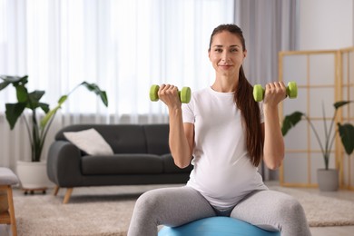 Beautiful pregnant woman with dumbbells doing exercises on fitball at home