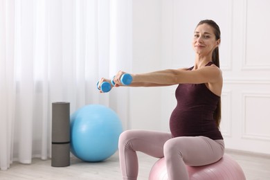 Beautiful pregnant woman with dumbbells doing exercises on fitball at home