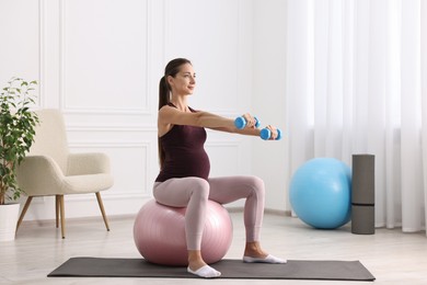 Beautiful pregnant woman with dumbbells doing exercises on fitball at home