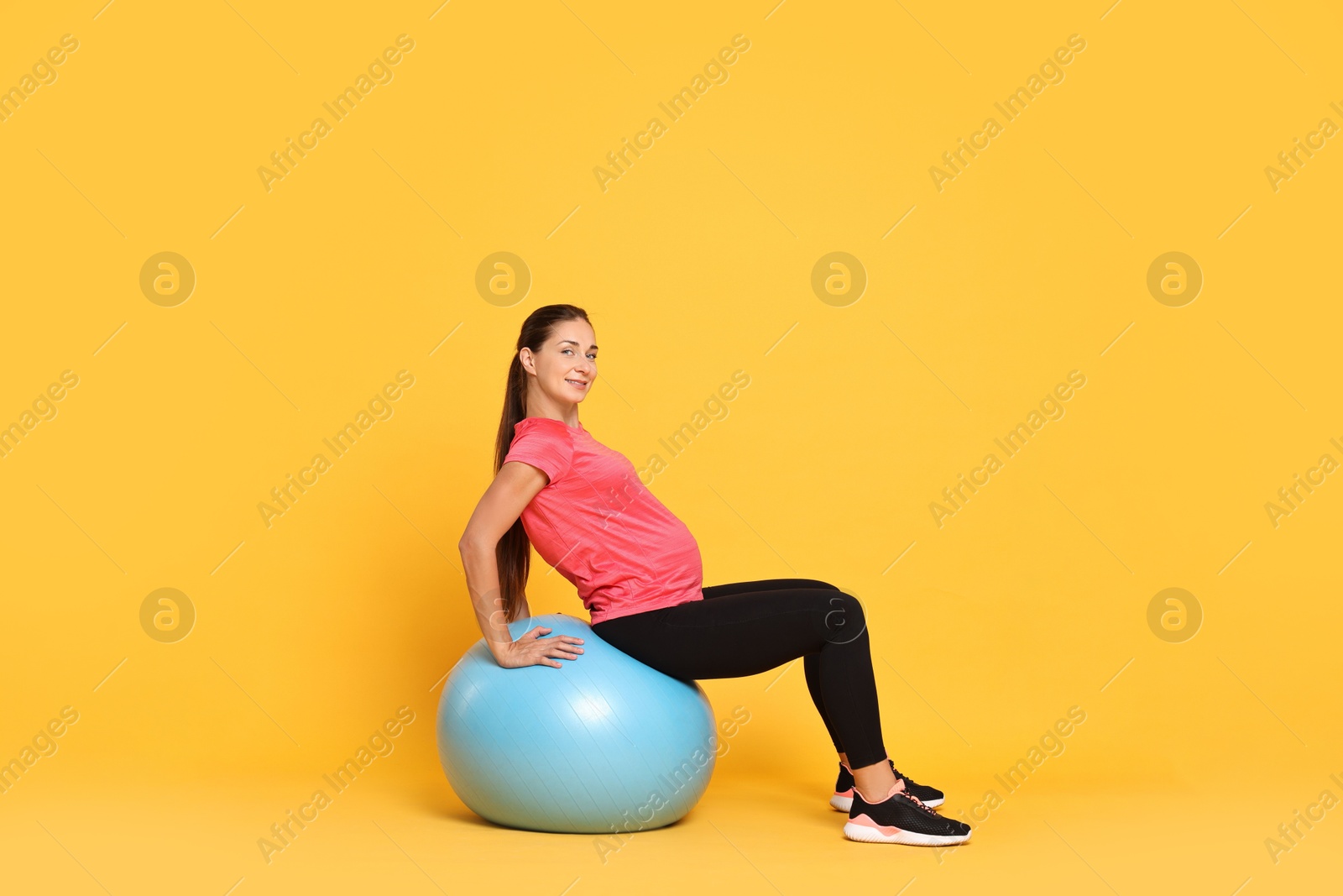 Photo of Beautiful pregnant woman doing exercises on fitball against yellow background