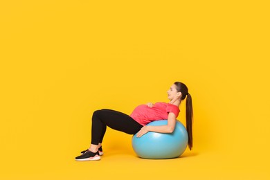 Beautiful pregnant woman doing exercises on fitball against yellow background