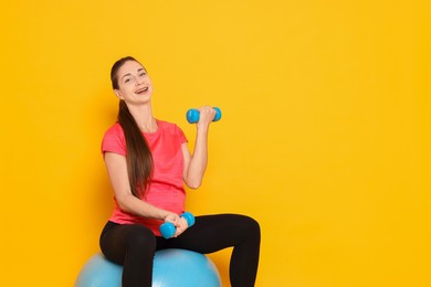 Beautiful pregnant woman with dumbbells doing exercises on fitball against yellow background, space for text