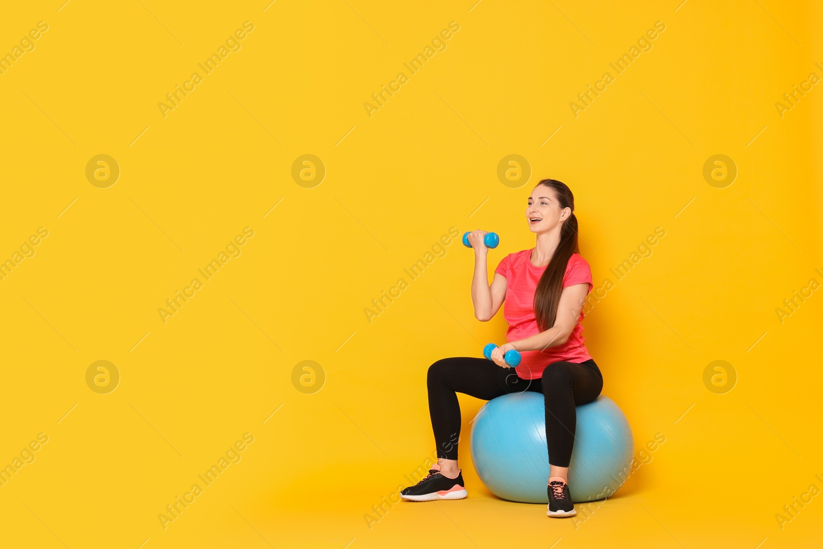 Photo of Beautiful pregnant woman with dumbbells doing exercises on fitball against yellow background, space for text