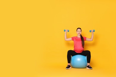 Photo of Beautiful pregnant woman with dumbbells doing exercises on fitball against yellow background, space for text