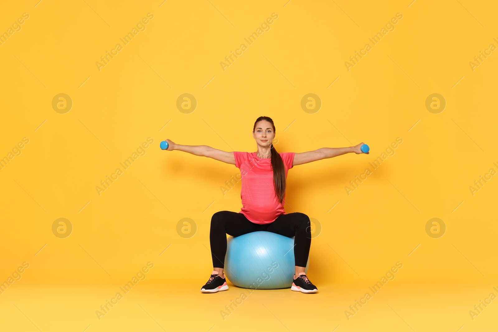 Photo of Beautiful pregnant woman with dumbbells doing exercises on fitball against yellow background