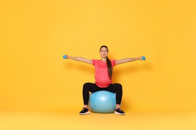 Beautiful pregnant woman with dumbbells doing exercises on fitball against yellow background