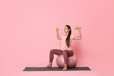 Photo of Beautiful pregnant woman with dumbbells doing exercises on fitball against pink background