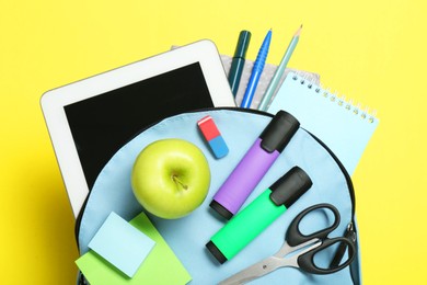 Photo of Light blue school backpack with stationery, apple and tablet on yellow background, top view