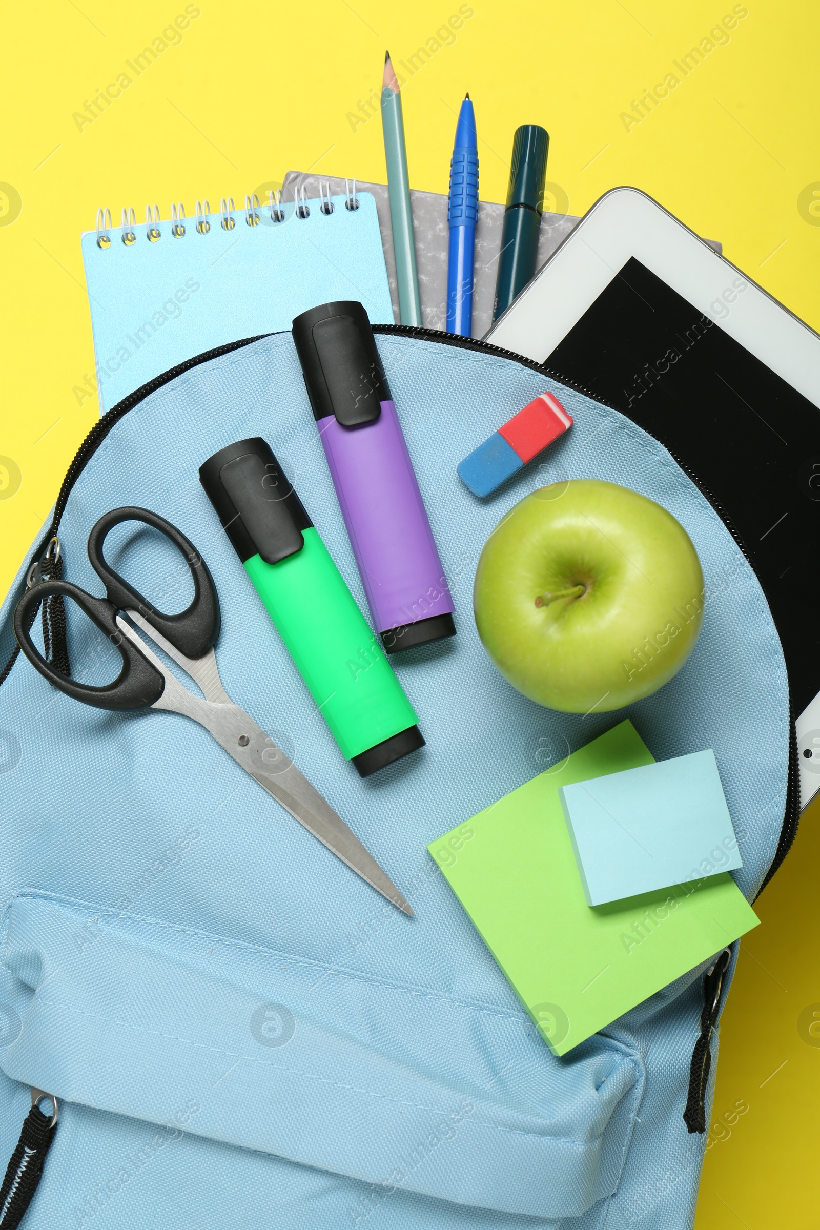 Photo of Light blue school backpack with stationery, apple and tablet on yellow background, top view