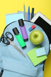 Light blue school backpack with stationery, apple and tablet on yellow background, top view