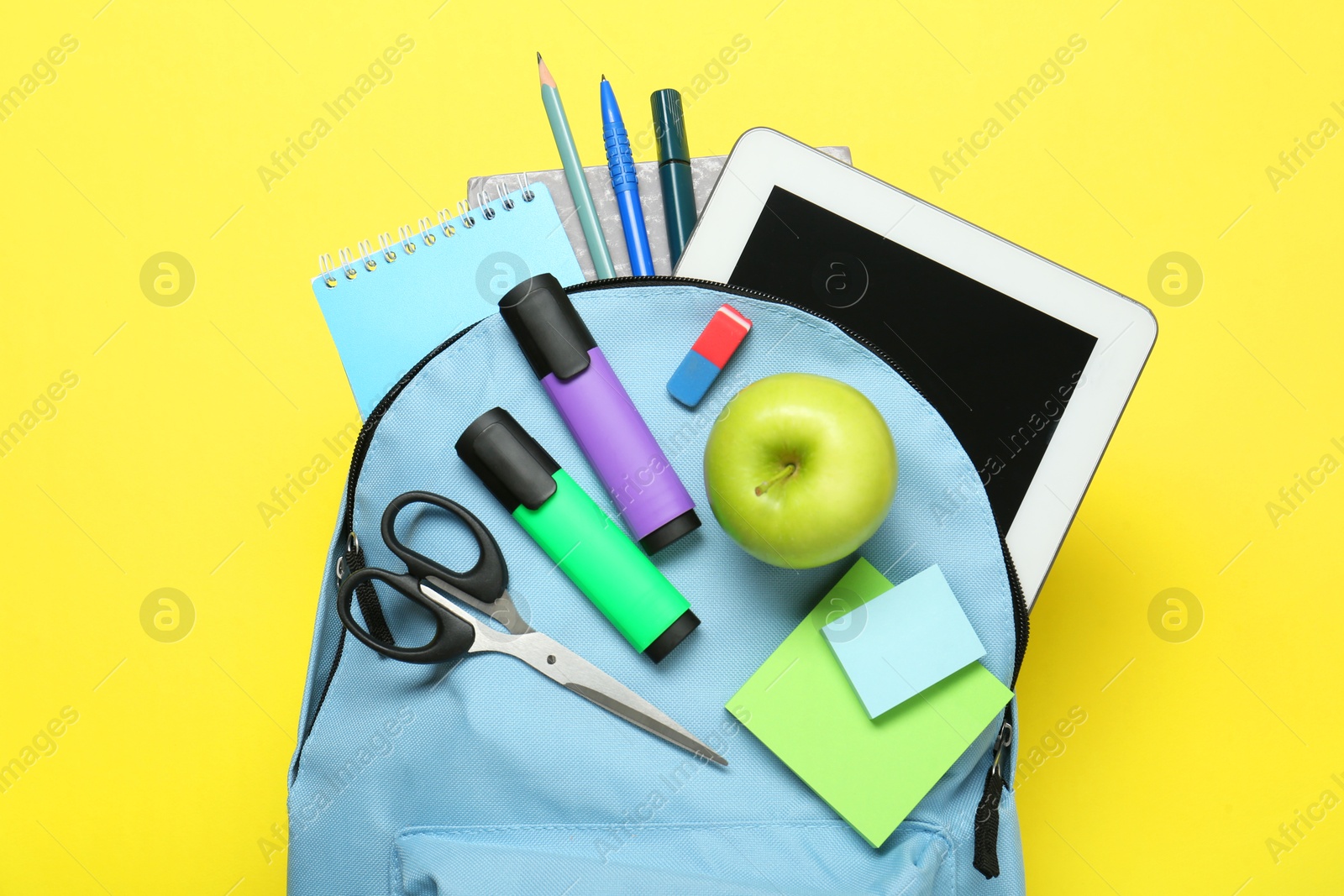 Photo of Light blue school backpack with stationery, apple and tablet on yellow background, top view